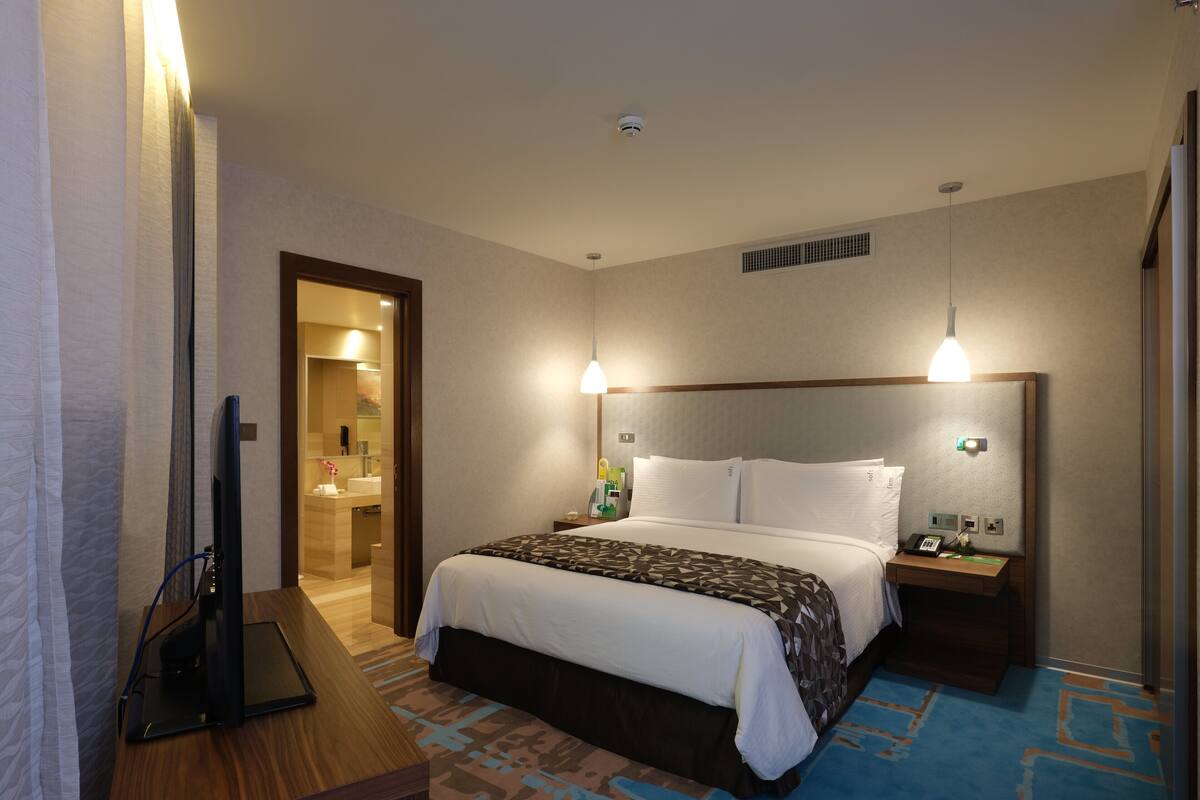 A hotel room with a bed, bedside tables, a desk with a computer, a bathroom with a mirror and sink, and a hallway.