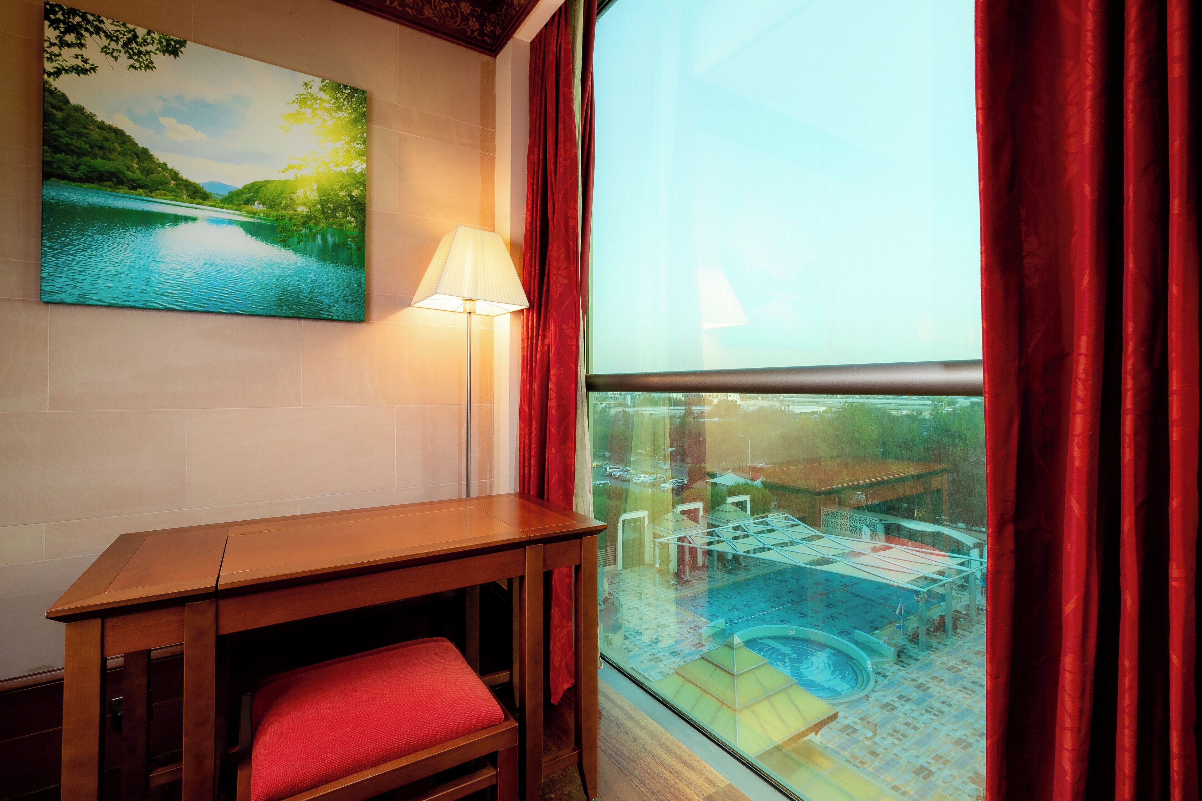 Una habitación con una mesa de madera, una silla roja, un cuadro de un paisaje y una ventana con vista a una piscina y zona exterior.