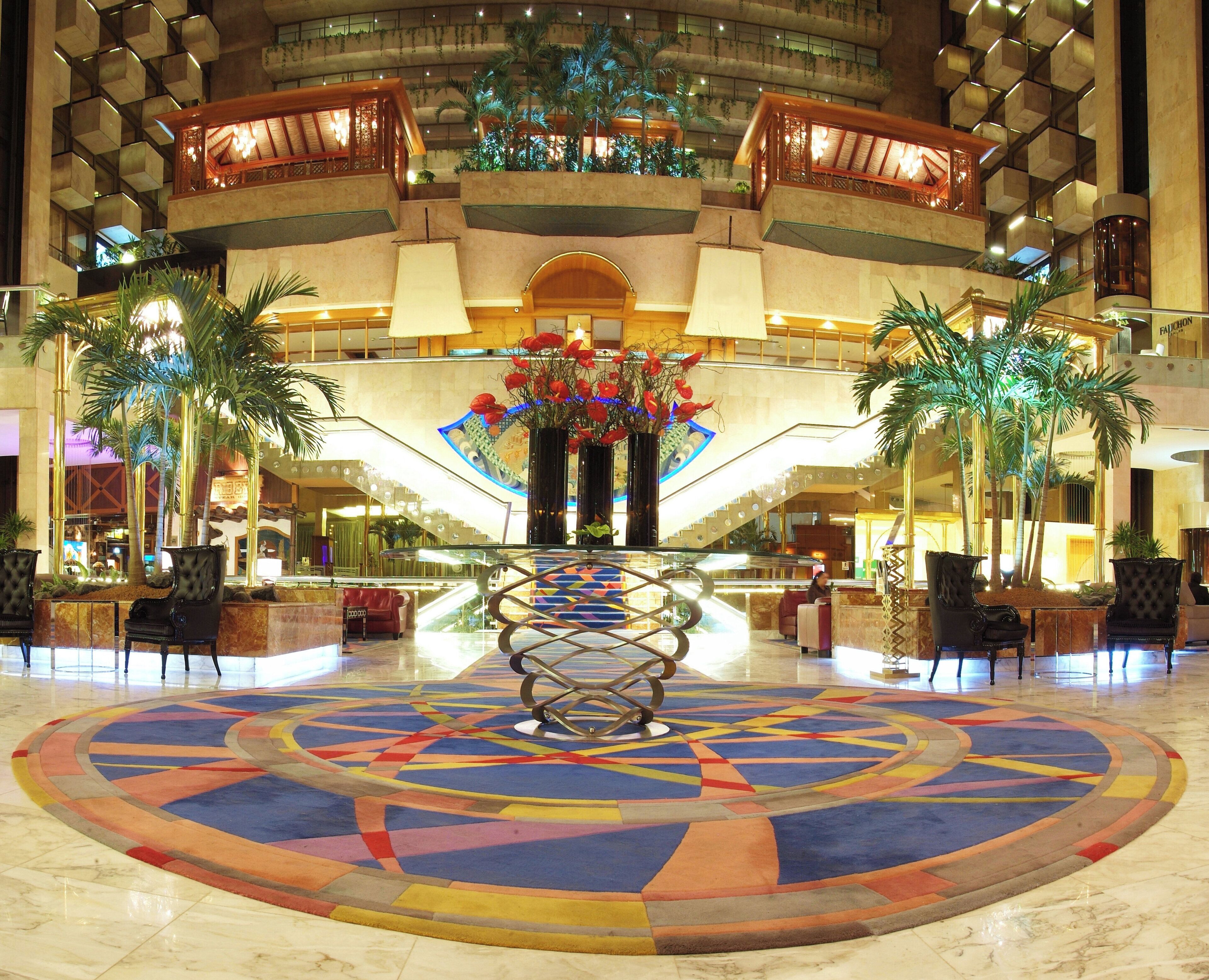 Un vestíbulo de hotel con una pieza decorativa central, varias zonas de estar y una escalera que conduce a plantas superiores.