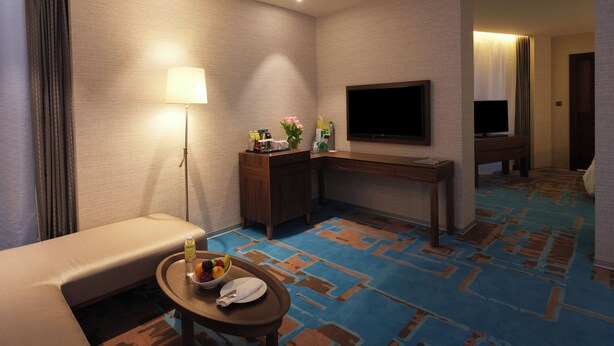 Une chambre d’hôtel avec un canapé, une petite table sur laquelle se trouve un saladier rempli de fruits et une bouteille, un bureau avec un téléviseur à écran plat et une moquette à motifs.