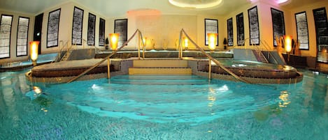 An indoor swimming pool with steps leading to a central area, surrounded by seating and decorative lighting.
