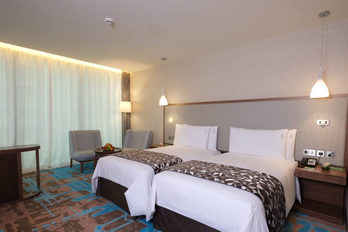 A hotel room with two beds, a desk, a chair, and a small table with fruit.