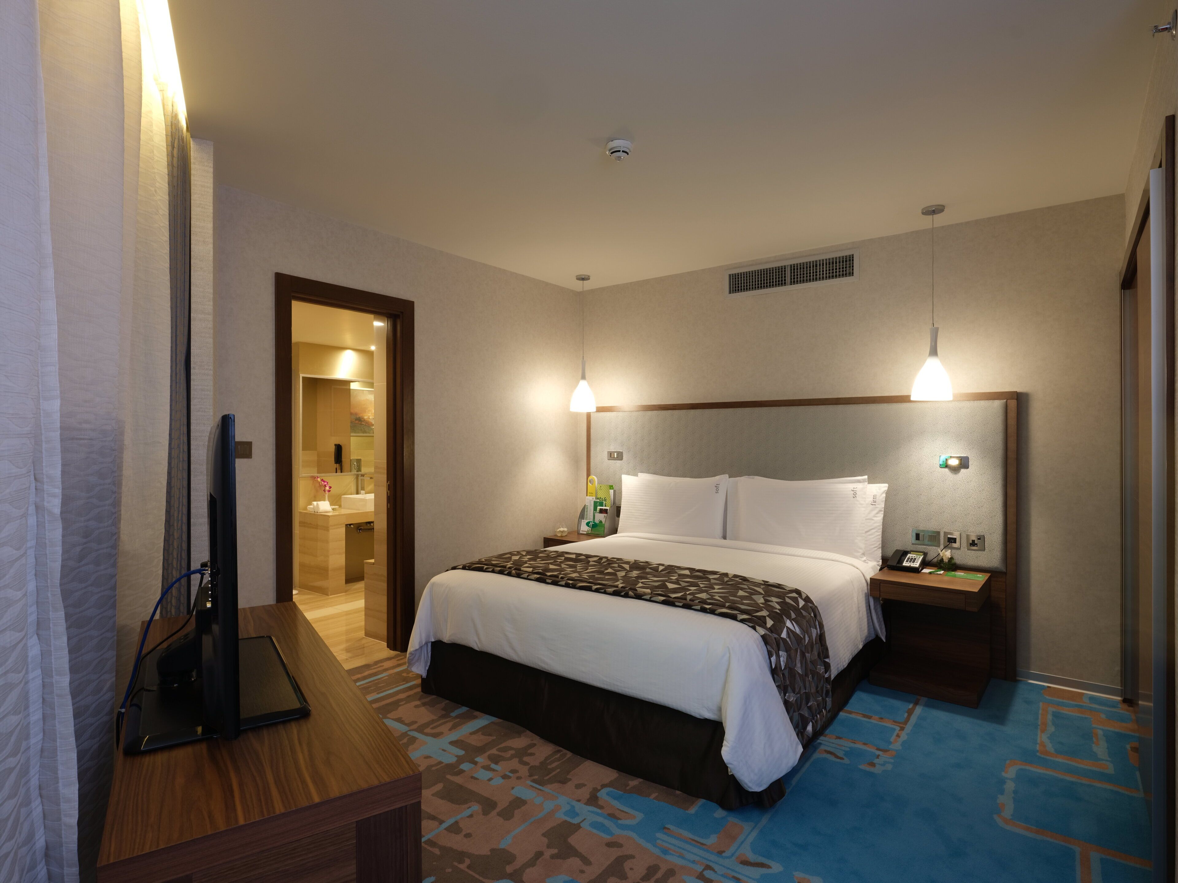 Ein Hotelzimmer mit Bett, Nachttischen, einem Schreibtisch mit Computer sowie einem Badezimmer mit Spiegel und Duschbereich.