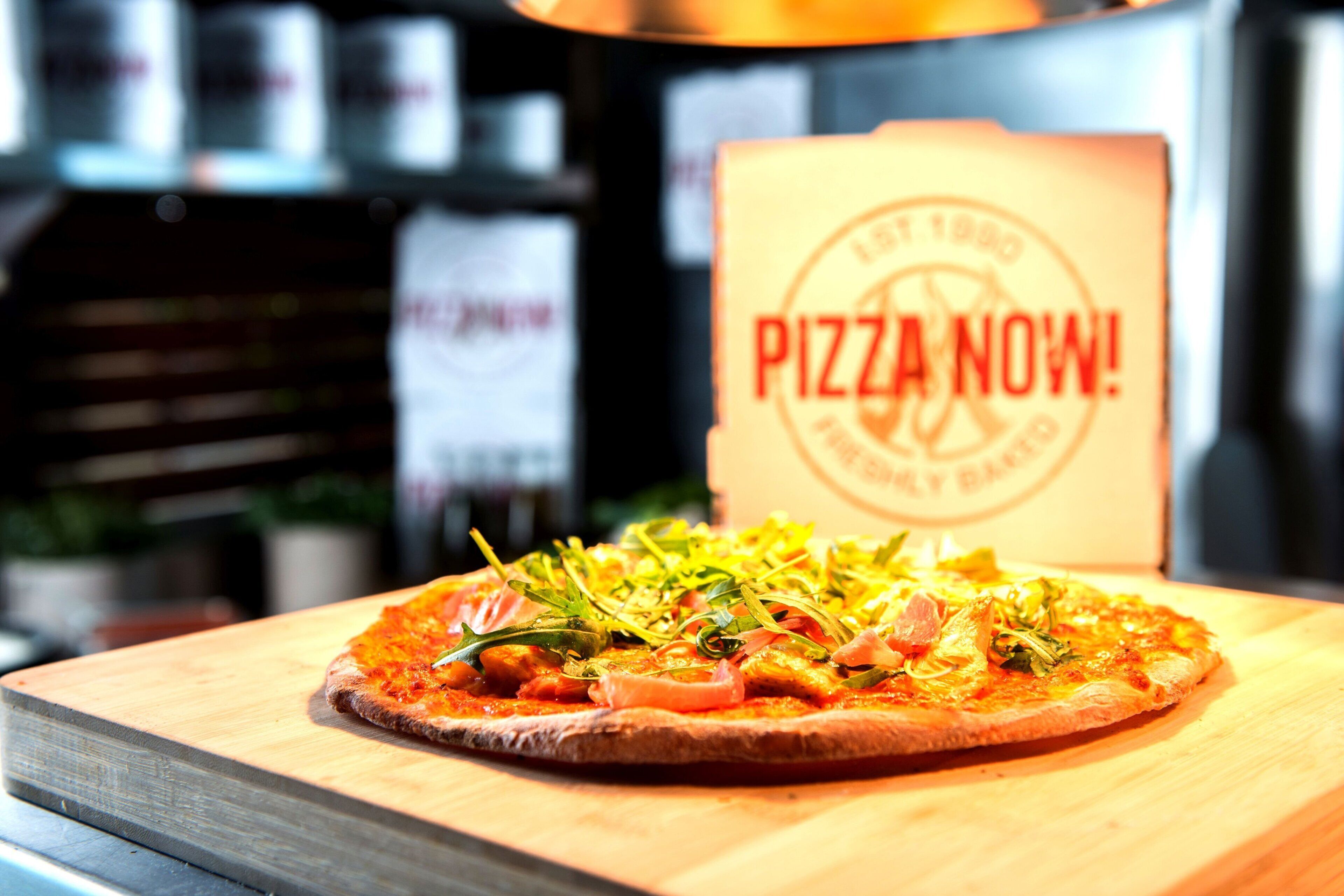 A pizza on a wooden board with a sign in the background that reads 'PIZZA NOW!'
