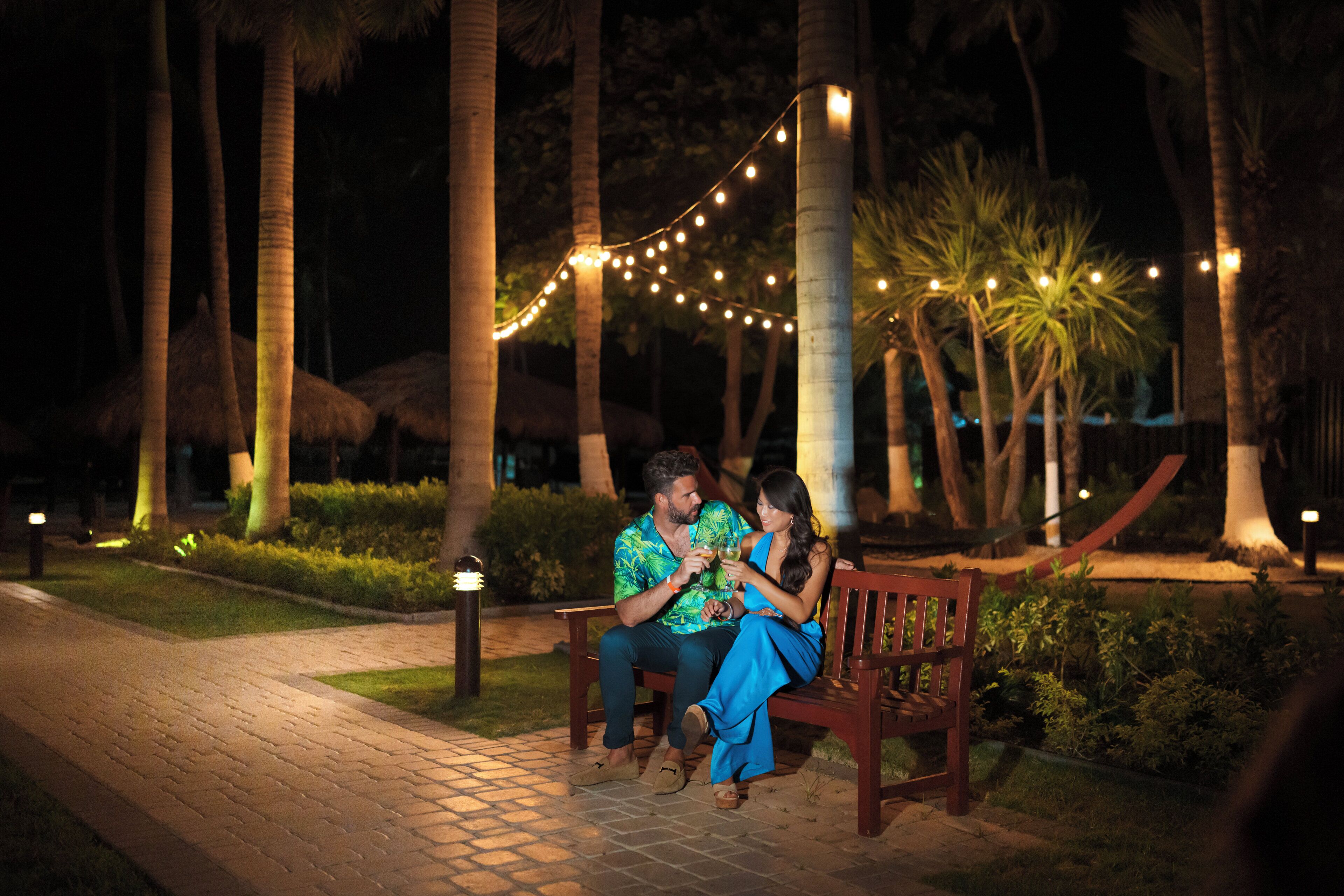 A couple seated on a bench in a well-lit outdoor area with string lights, surrounded by palm trees and a paved walkway.