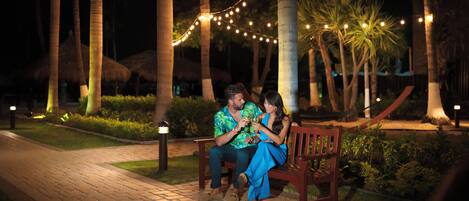 A couple seated on a bench in a well-lit outdoor area with string lights, surrounded by palm trees and a paved walkway.