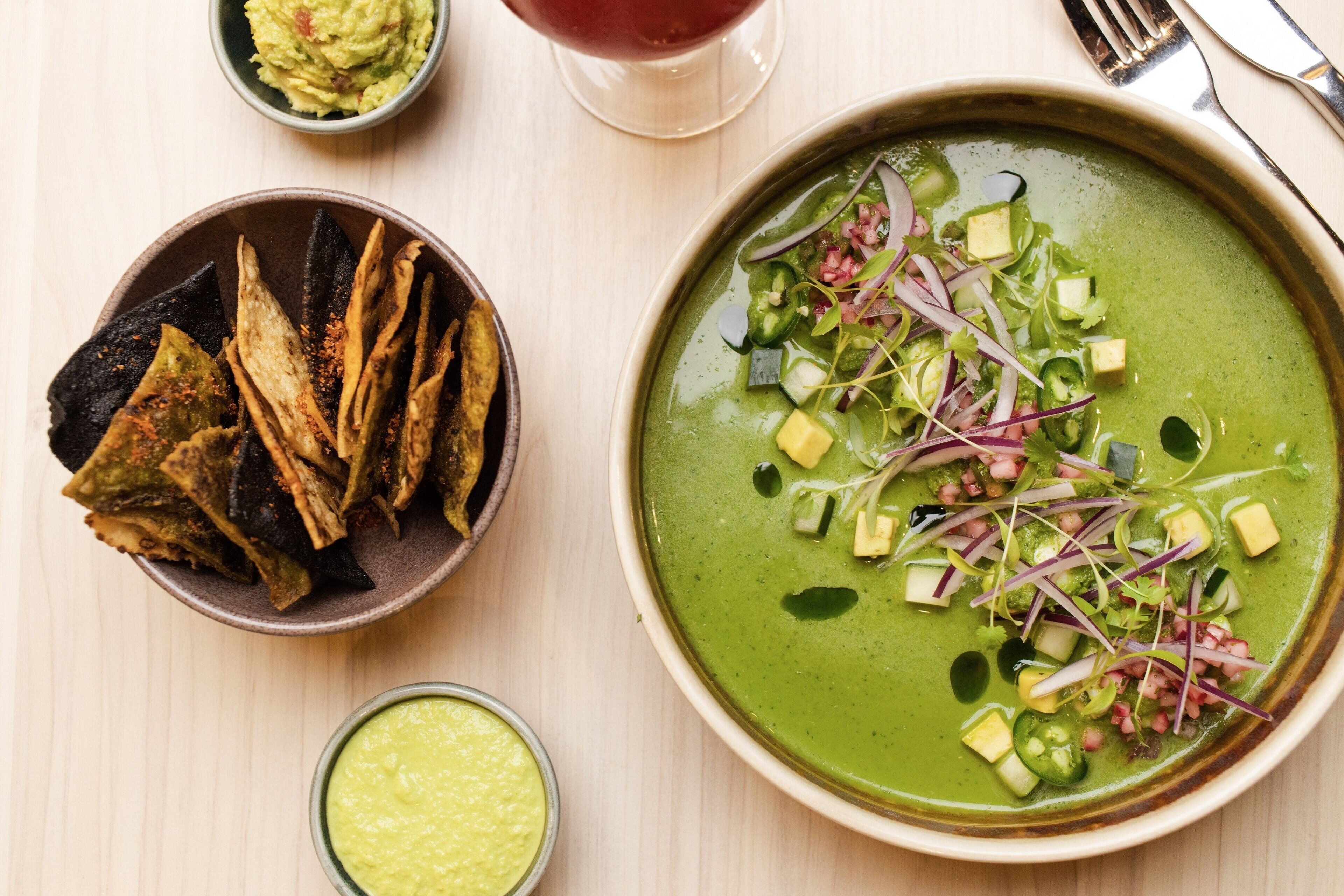A bowl of green soup garnished with vegetables and cheese, a bowl of tortilla chips, and a glass of red drink on a wooden table.