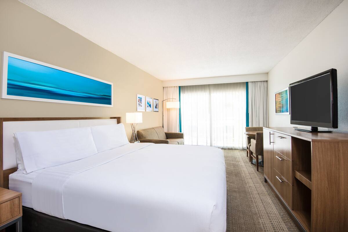A hotel room with a large bed, a television on a wooden cabinet, a desk with a chair, and a large window with curtains.