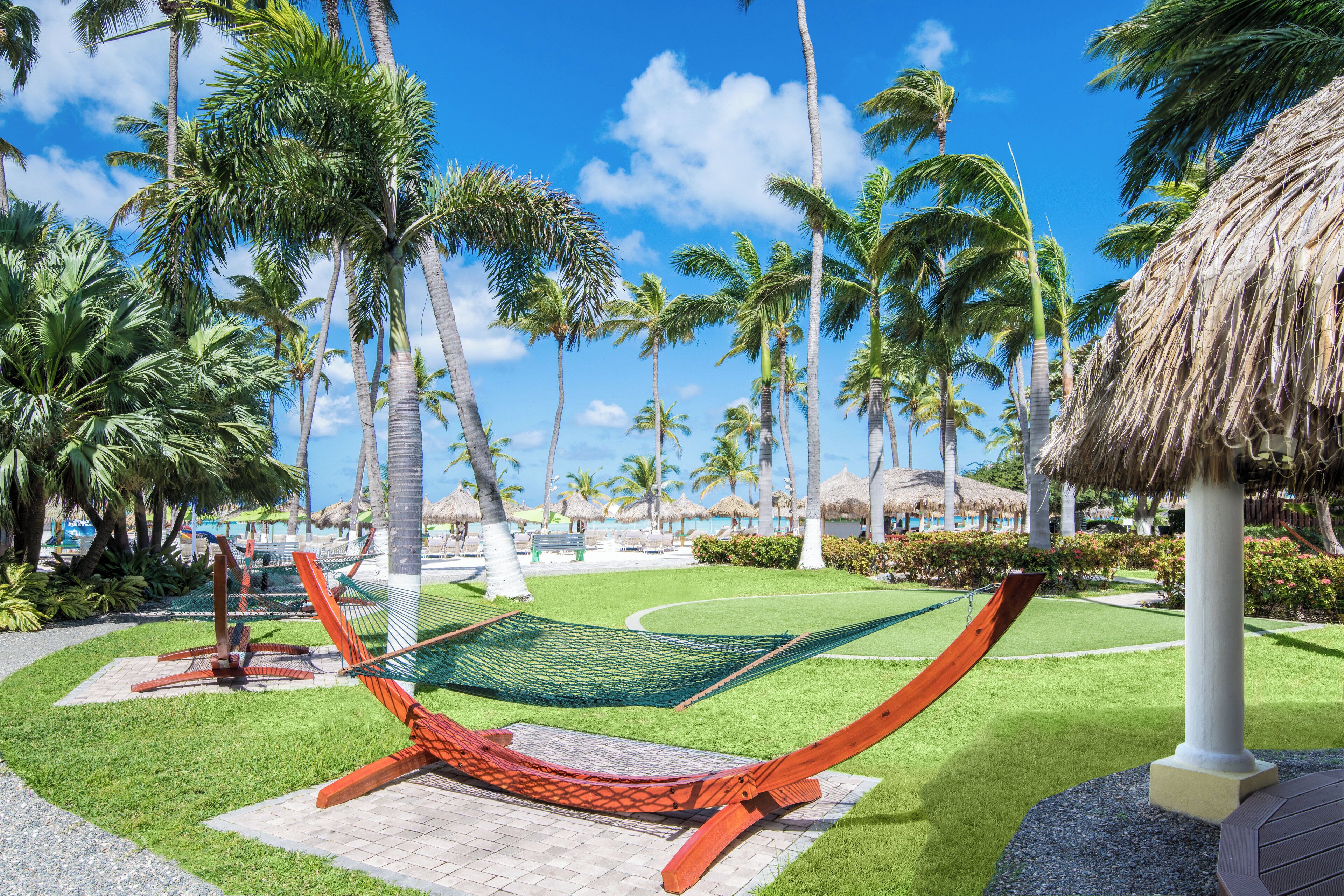 A hammock in a tropical garden with palm trees and a thatched-roof structure.