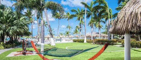 A hammock in a tropical garden with palm trees and a thatched-roof structure.