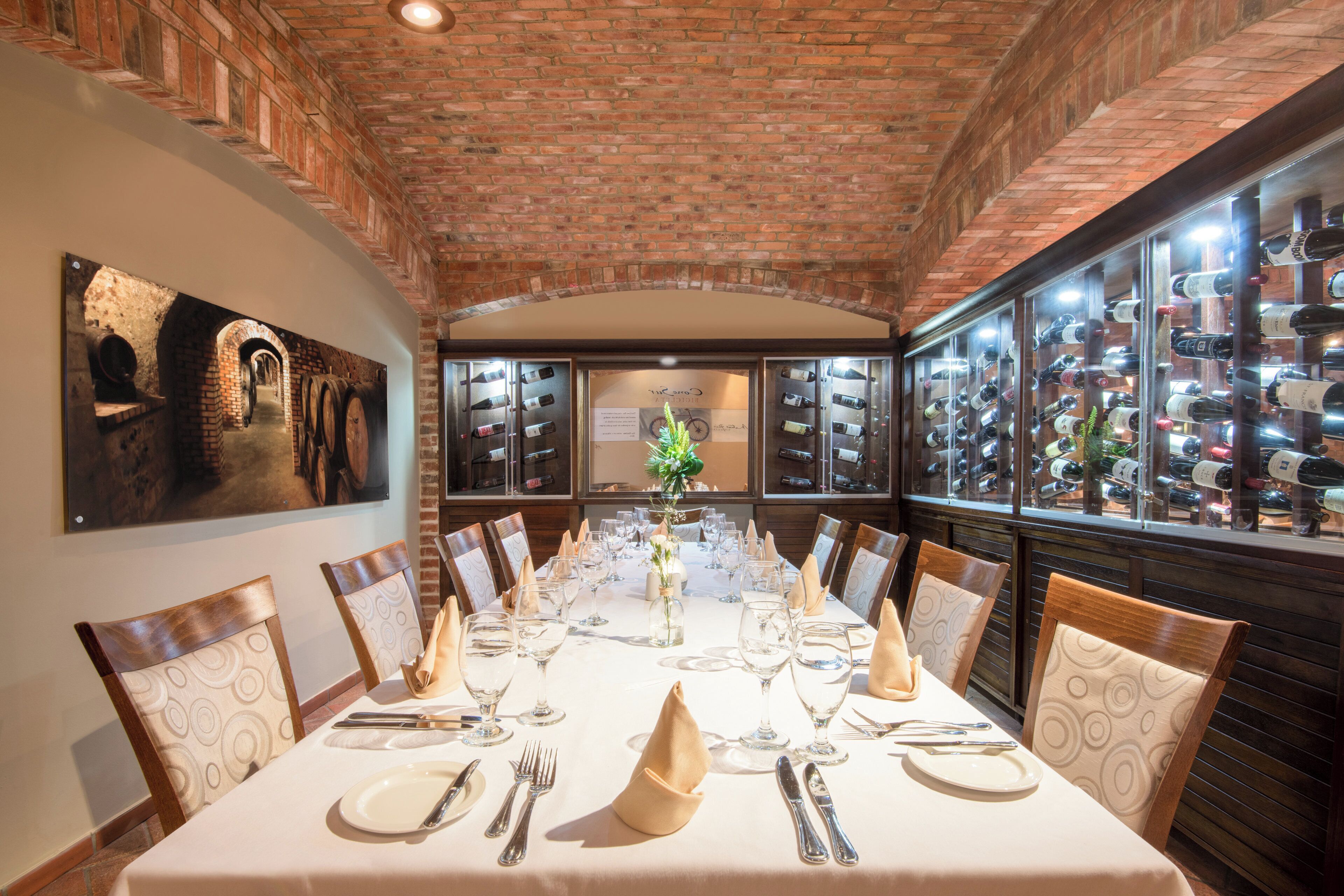 A long dining table set for a meal in a brick-walled room with wine bottles on display.