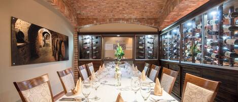 A long dining table set for a meal in a brick-walled room with wine bottles on display.
