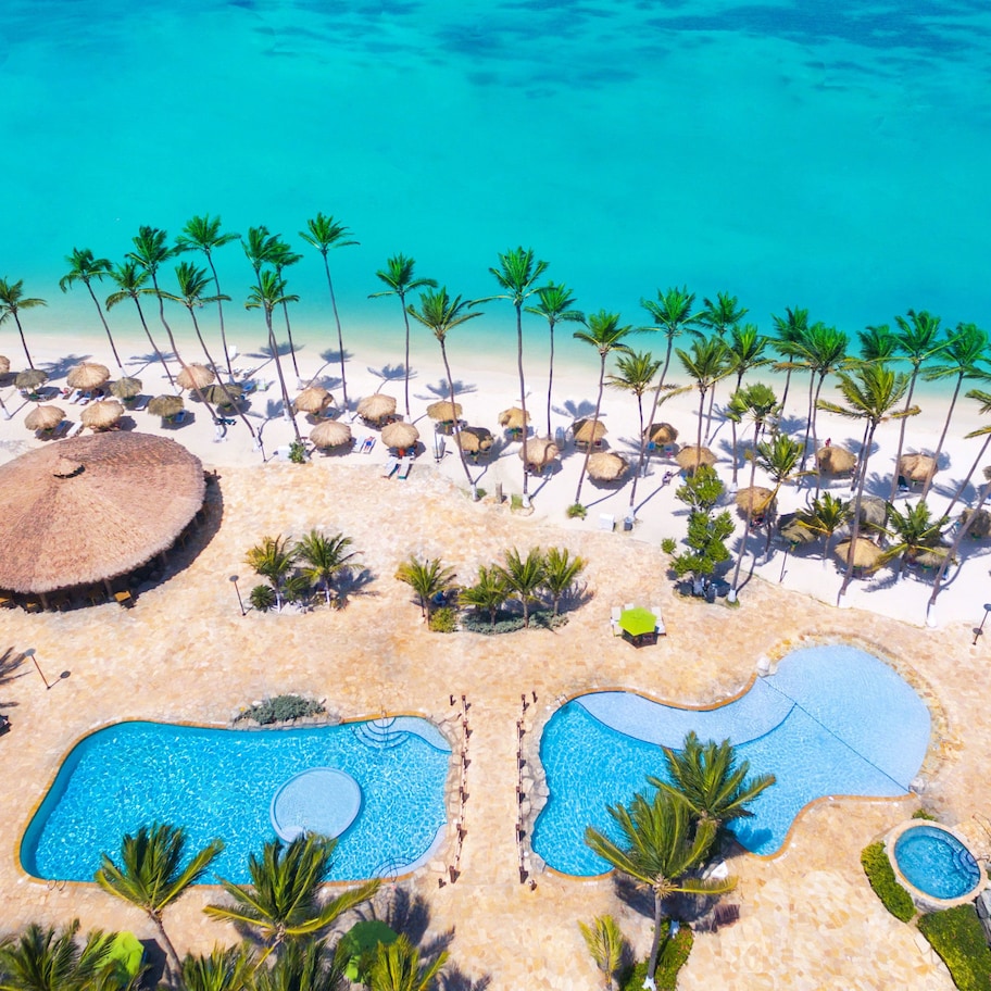 An aerial view of a beach resort with multiple swimming pools, palm trees, and thatched-roof huts.