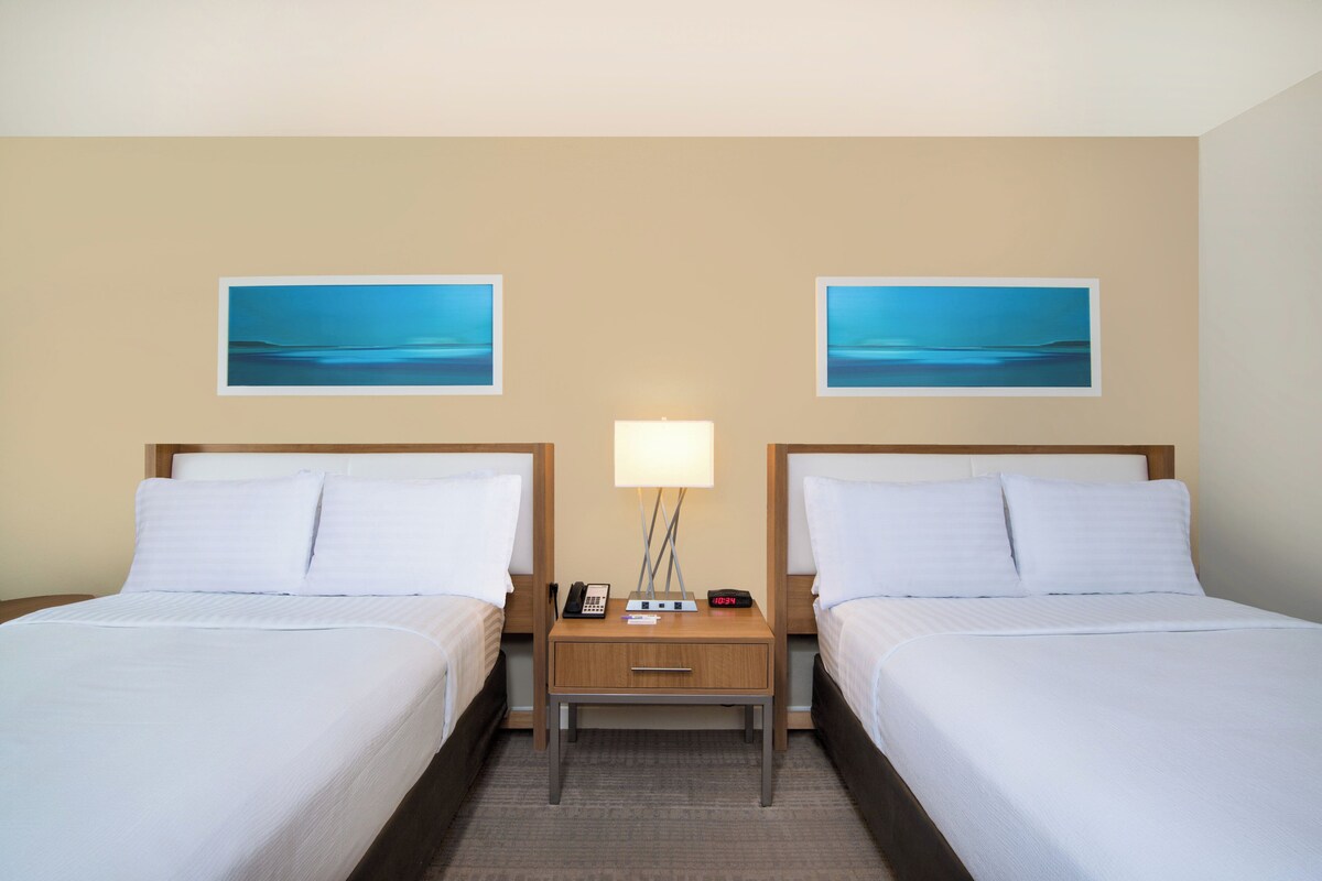 Una habitación de hotel con dos camas, una mesita de noche con lámpara, teléfono y reloj digital.