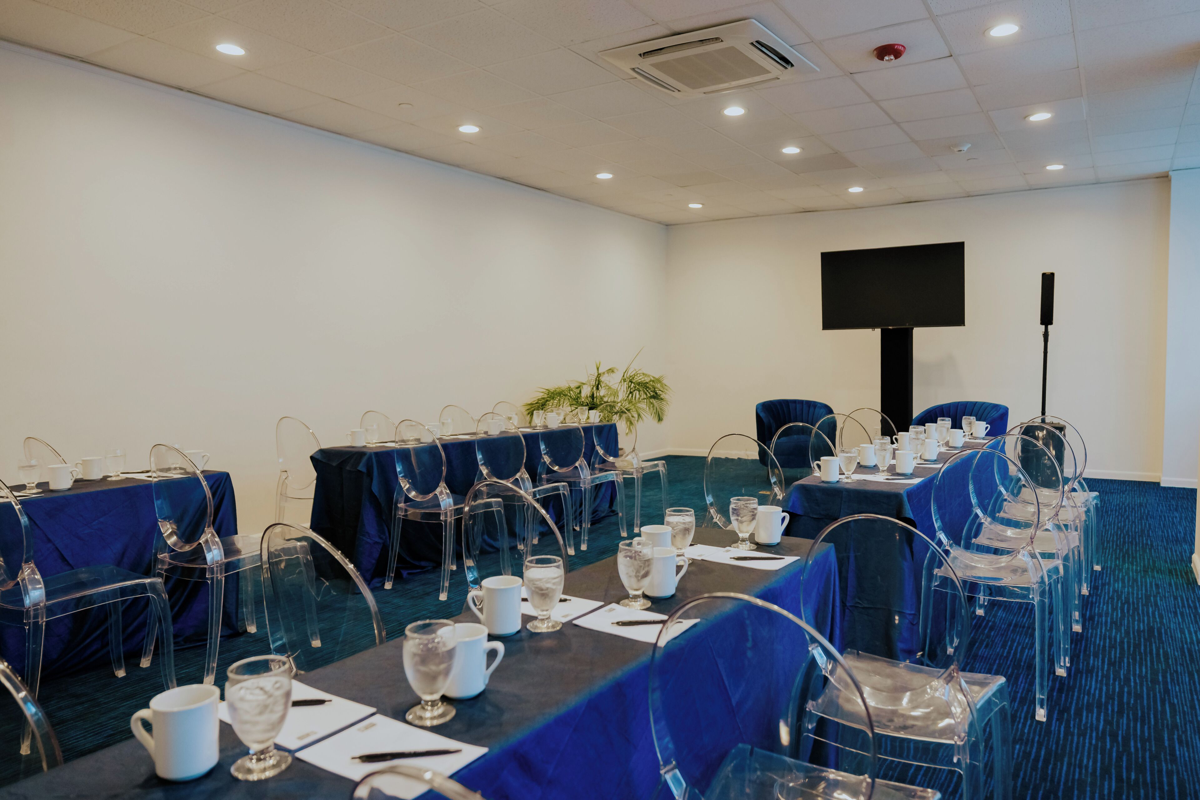A conference room with tables set for a meeting, clear chairs, a large screen, and a plant.