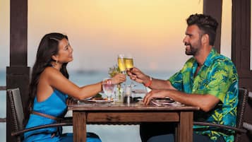 A couple toasting with drinks at a wooden table with a view of the ocean.