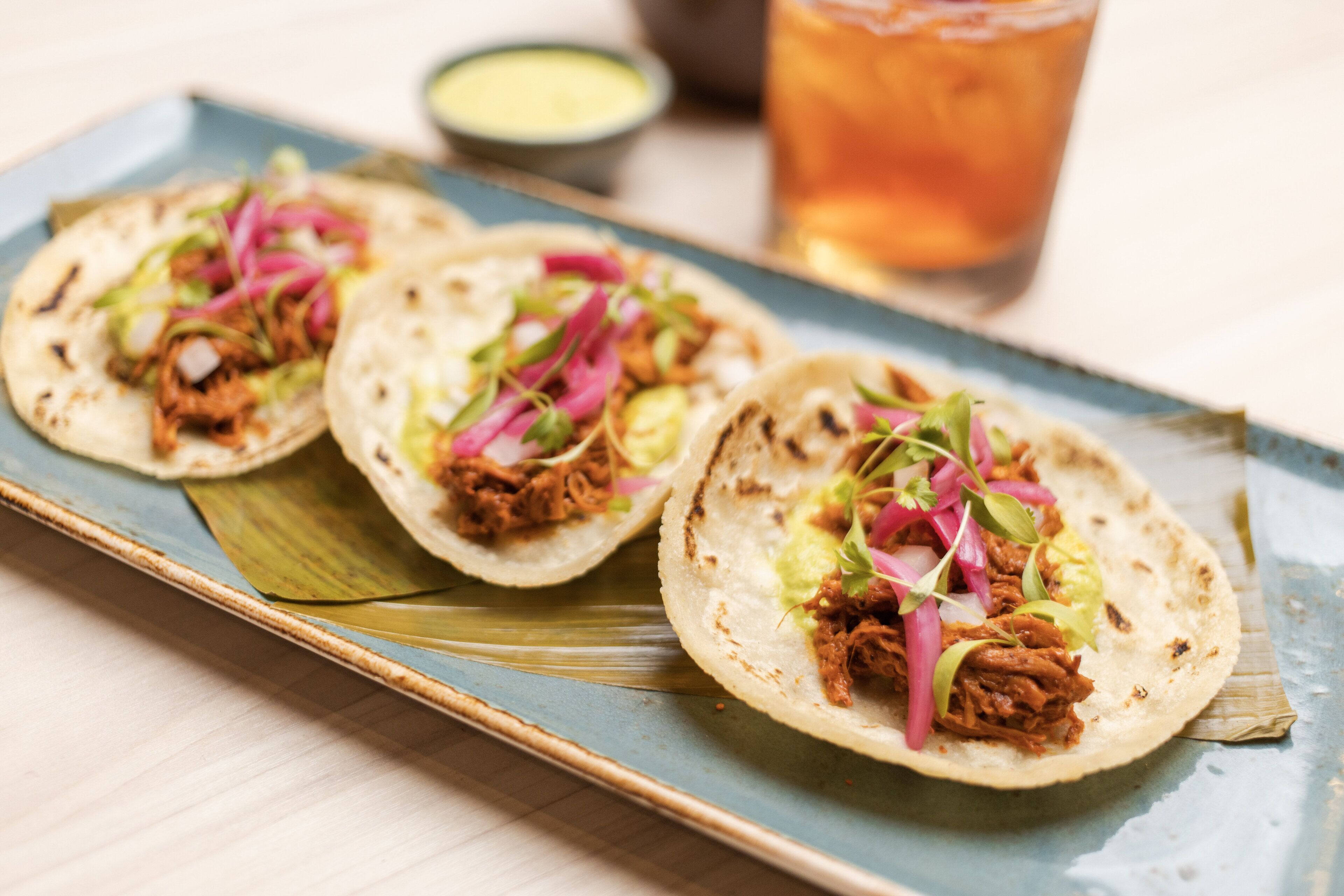 Three tacos on a blue plate with a drink in the background.