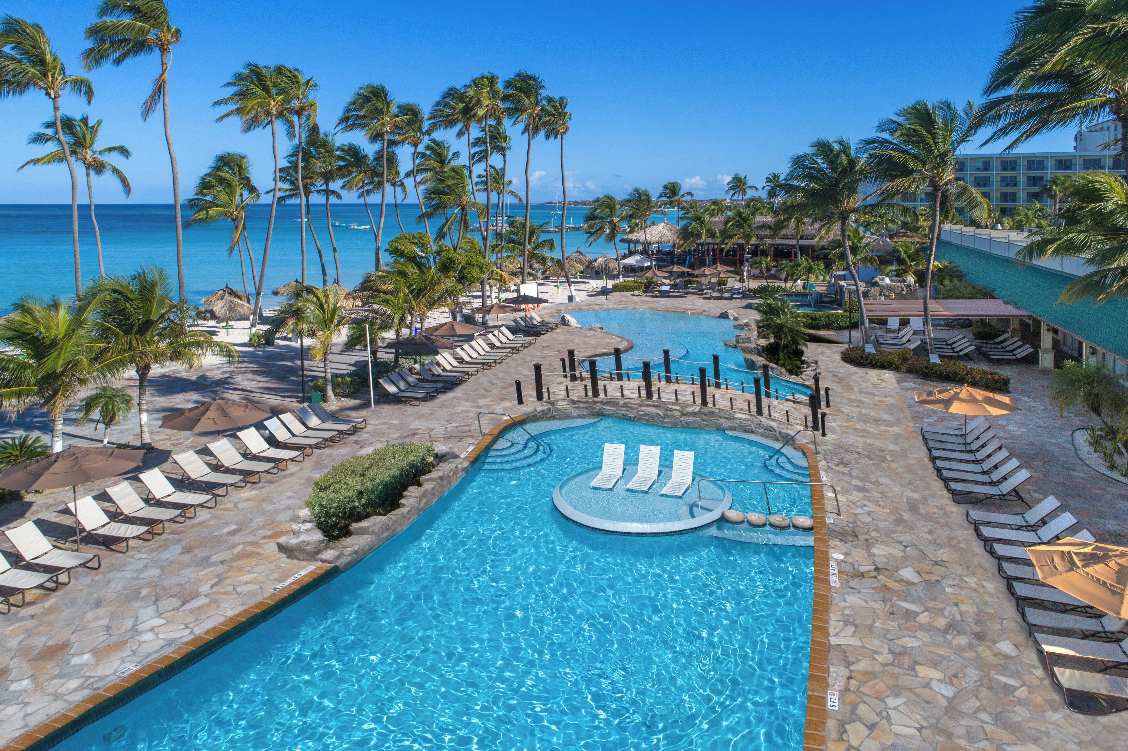 A resort pool area with multiple swimming pools, sun loungers, and palm trees.