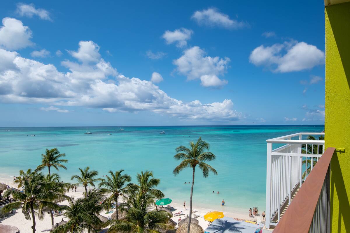 A beach view with clear turquoise water, palm trees, and a white building with a balcony.