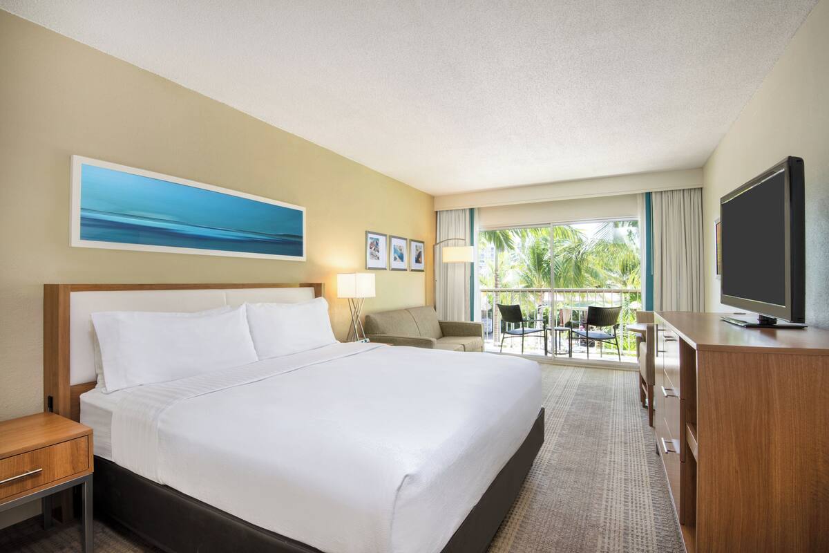 A hotel room with a large bed, a desk, a television, and a balcony with outdoor seating.
