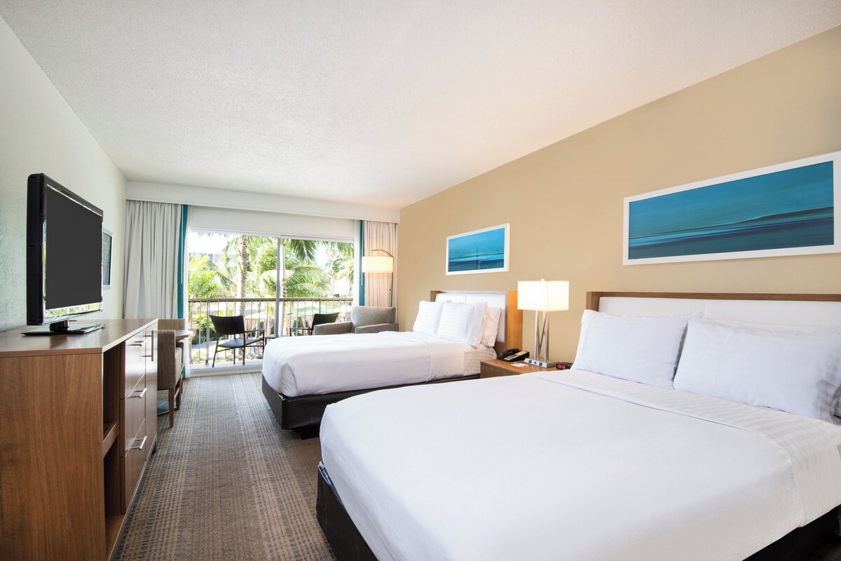 Habitación de hotel con dos camas, un escritorio, un televisor y un balcón con vista a palmeras.