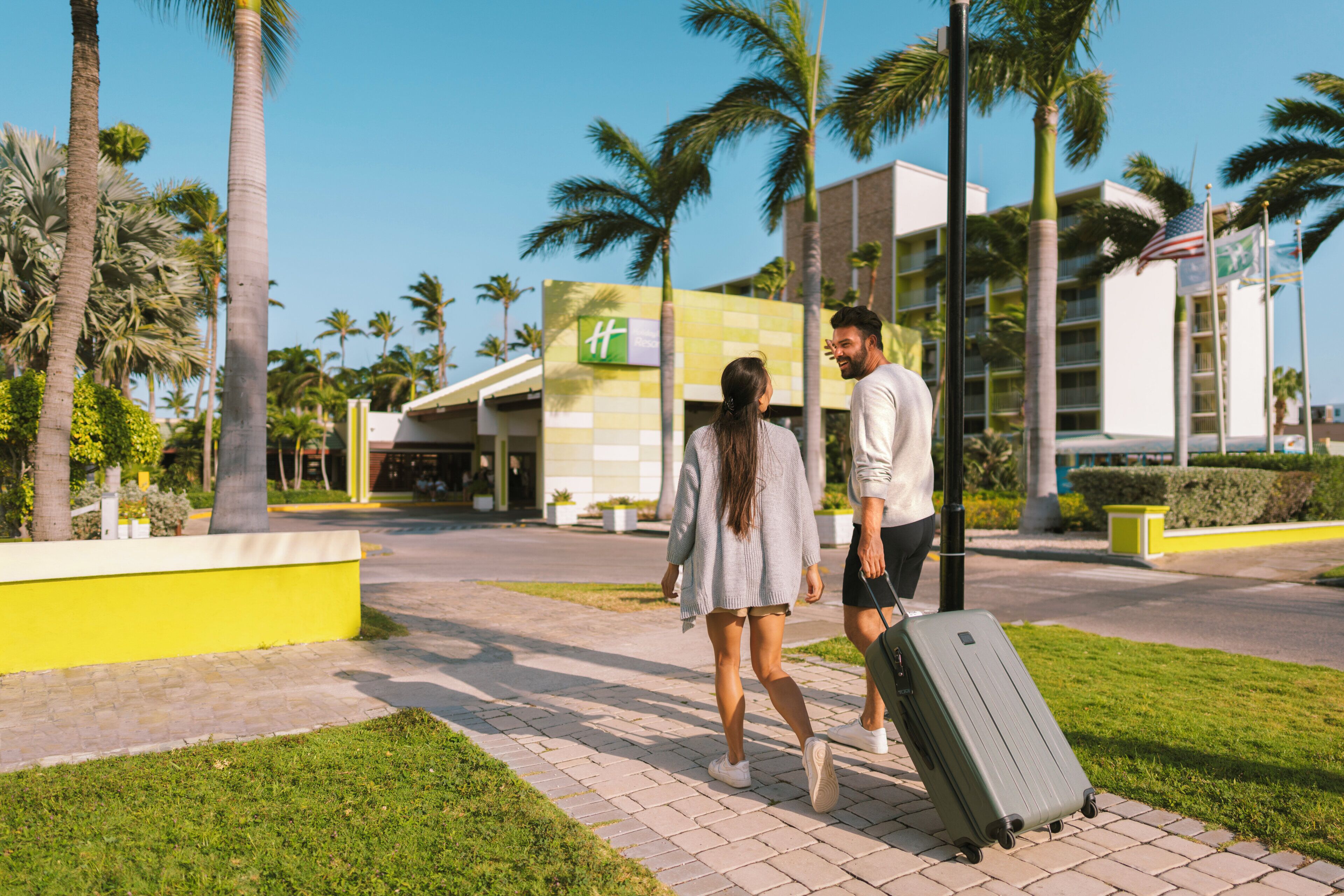 A couple walking on a paved pathway with a large suitcase, palm trees, and a building with a green and yellow facade in the background.
