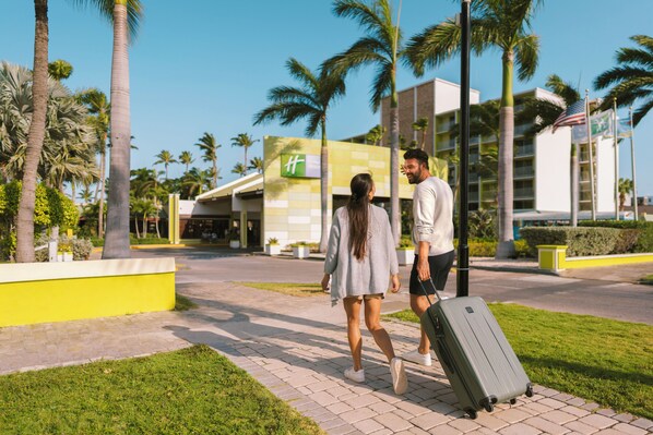 Una pareja caminando por un sendero pavimentado con una maleta grande, palmeras y un edificio con fachada verde y amarilla al fondo.