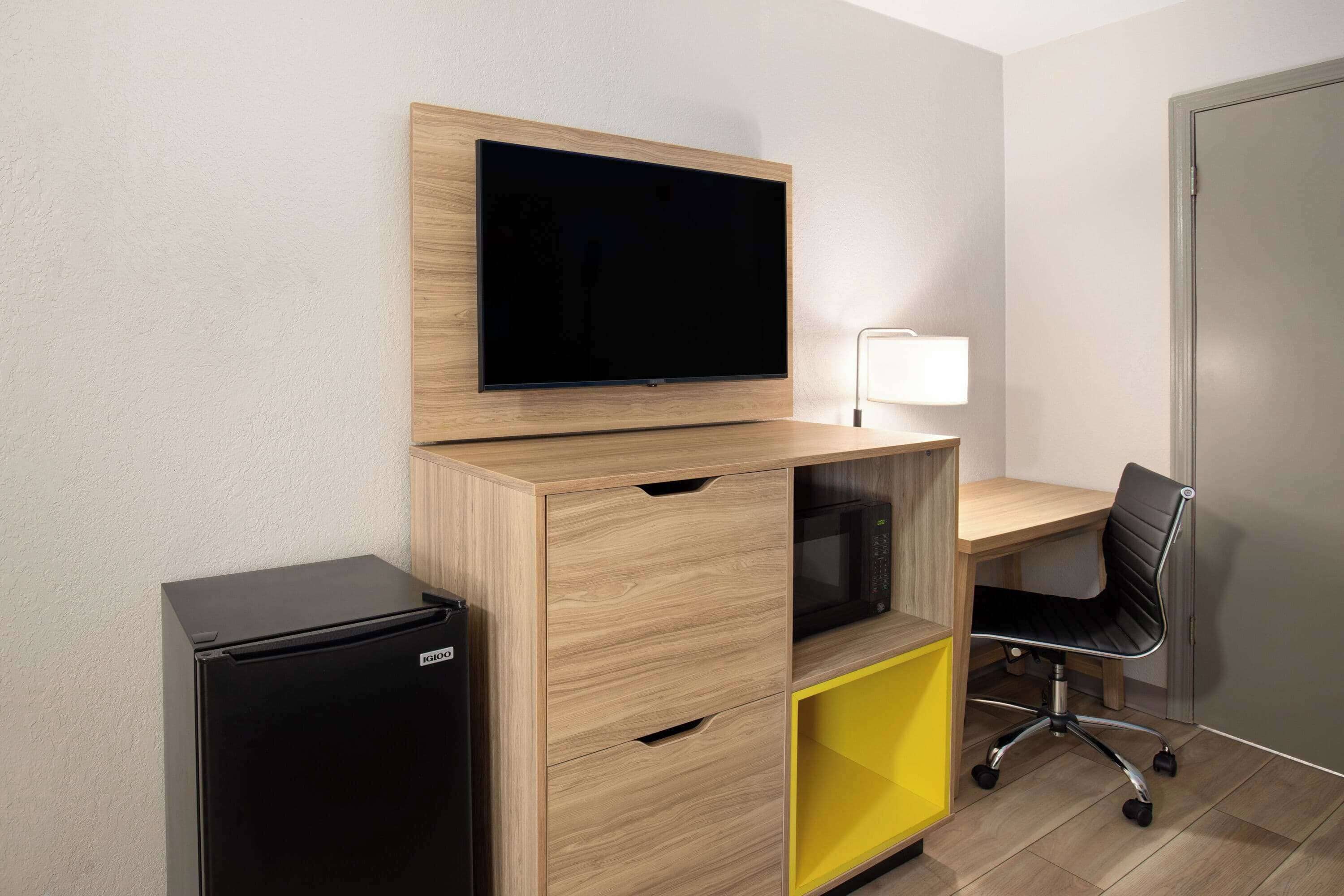 Una habitación compacta con un escritorio de madera, una silla de oficina negra, un televisor de pantalla plana en la pared y un refrigerador negro pequeño.