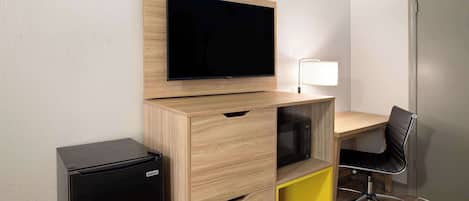 A compact room with a wooden desk, a black office chair, a flat-screen TV mounted on the wall, and a small black refrigerator.