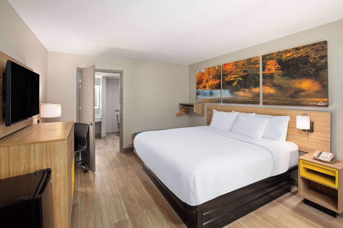 Quarto de hotel com uma cama grande, uma escrivaninha com televisão de tela plana, uma poltrona e uma luminária de mesa. Há duas pinturas na parede acima da cama.