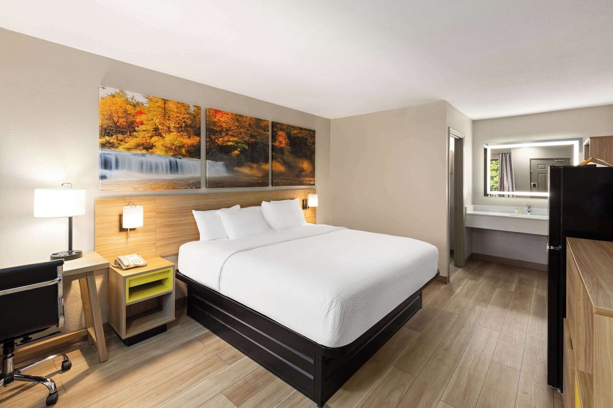 Quarto de hotel com cama, escrivaninha, cadeira, mesa de cabeceira e uma janela grande.
