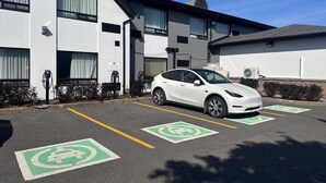 Un coche blanco estacionado en un espacio reservado para vehículos eléctricos, con un edificio al fondo.