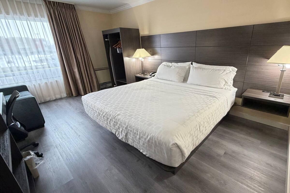 A hotel room with a large bed, a desk with a chair, a wardrobe, and a nightstand with a lamp.