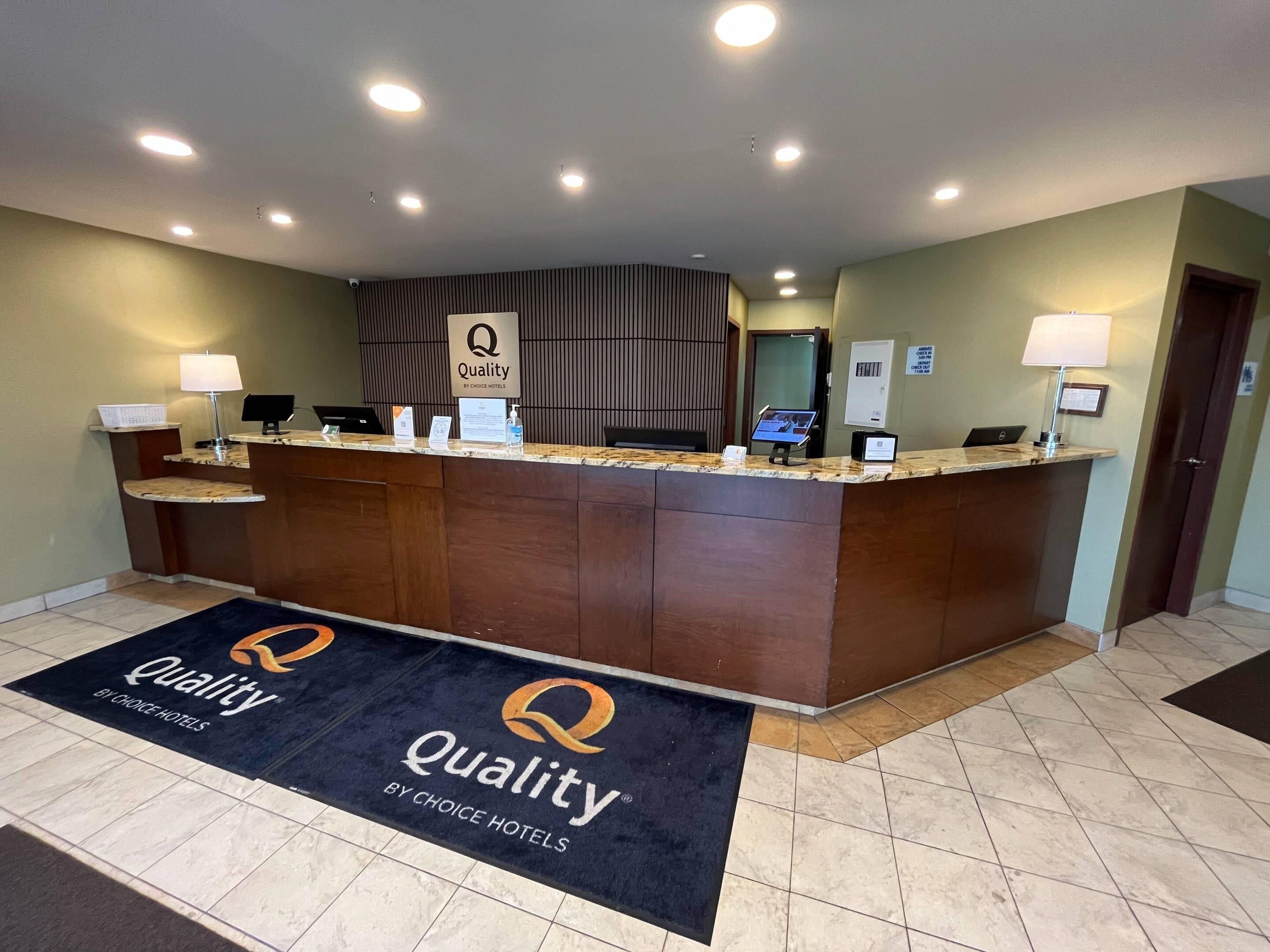 A hotel reception desk with a dark wood finish, a 'Quality by Choice Hotels' sign, a reception counter with a computer and a phone, and a black doormat with the hotel logo.