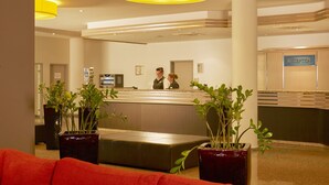 A reception area with a reception desk, two receptionists, a sign reading "RECEPTION", and potted plants. - Sunday Hotel Leipzig-Halle (Landsberg)