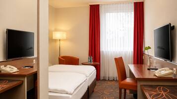 A hotel room with a bed, a desk with a telephone, a chair, a TV, and a window with curtains.