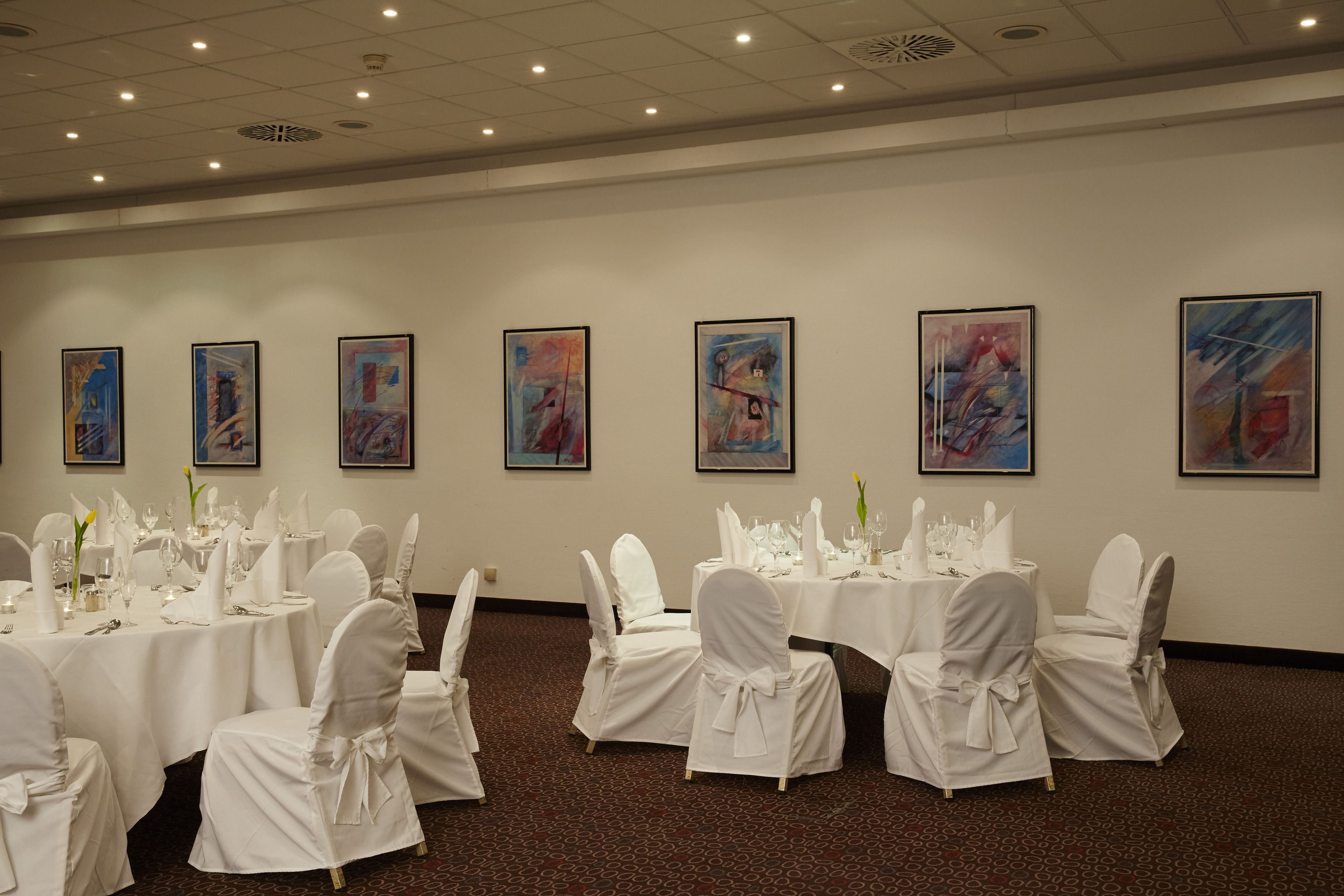 A banquet hall with tables set for a formal event, white chairs, and abstract paintings on the walls.