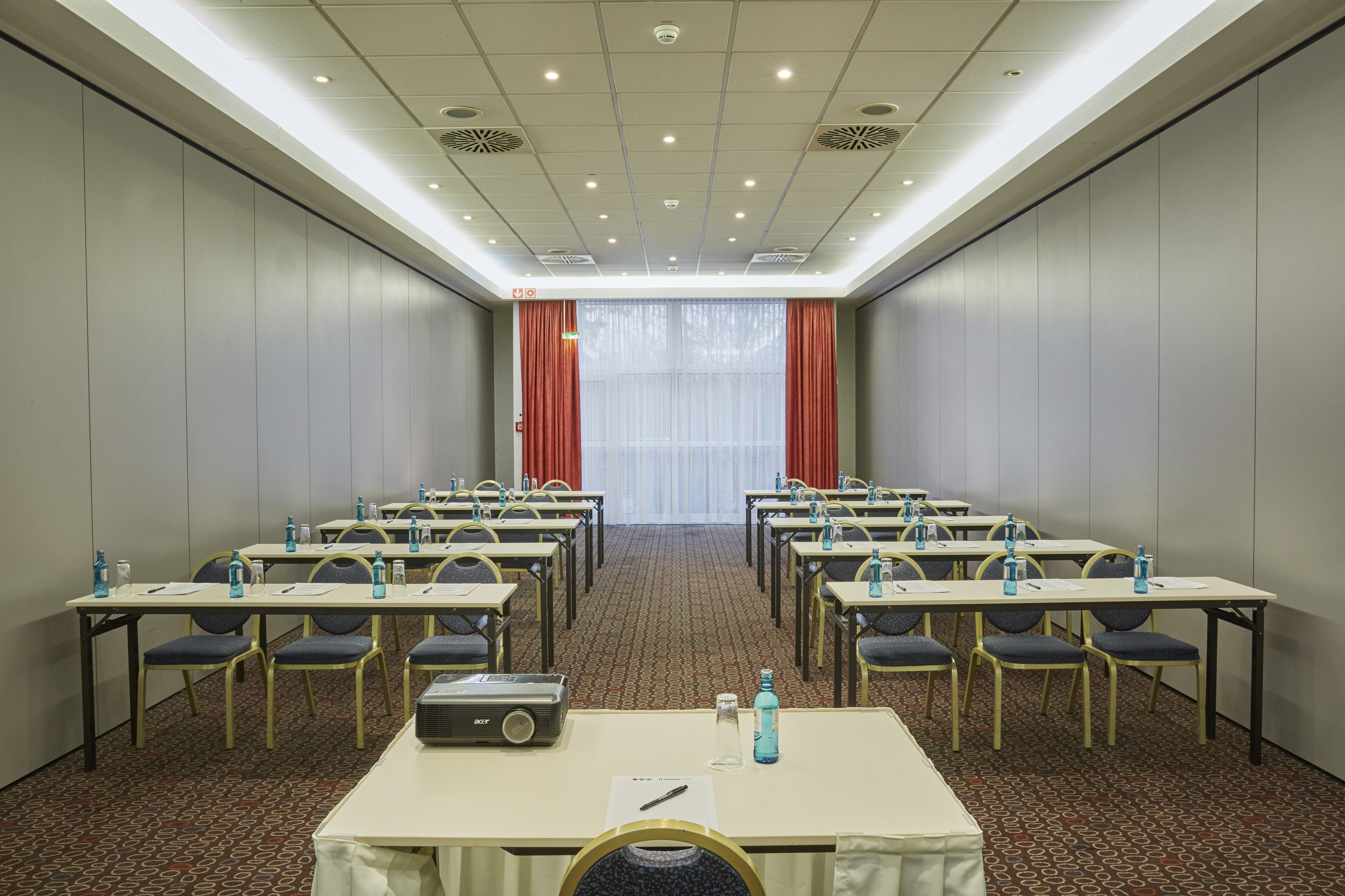 Un salón de conferencias con filas de mesas y sillas, equipado con botellas de agua y un proyector.
