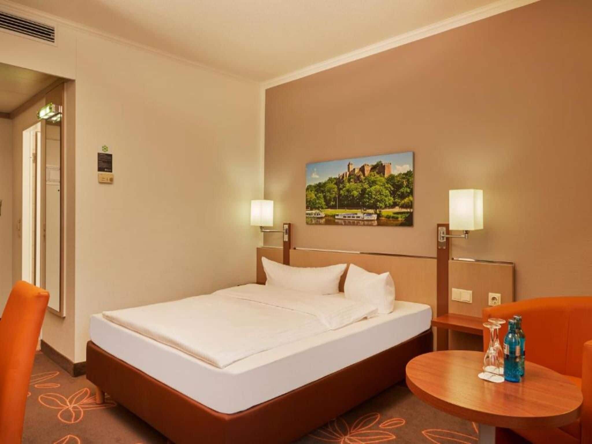 Een hotelkamer met een bed, twee nachtkastjes met lampen, een klein rond tafeltje met een fles en glazen, en een schilderij aan de muur.
