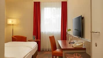Habitación de hotel con cama, escritorio con teléfono, una silla, televisor y ventana con cortinas.