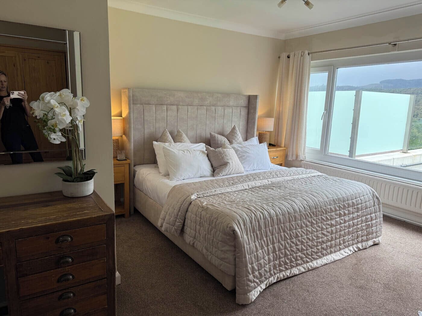 A bedroom with a large bed, a nightstand, a dresser, a potted orchid, and a view of the sea through a window.