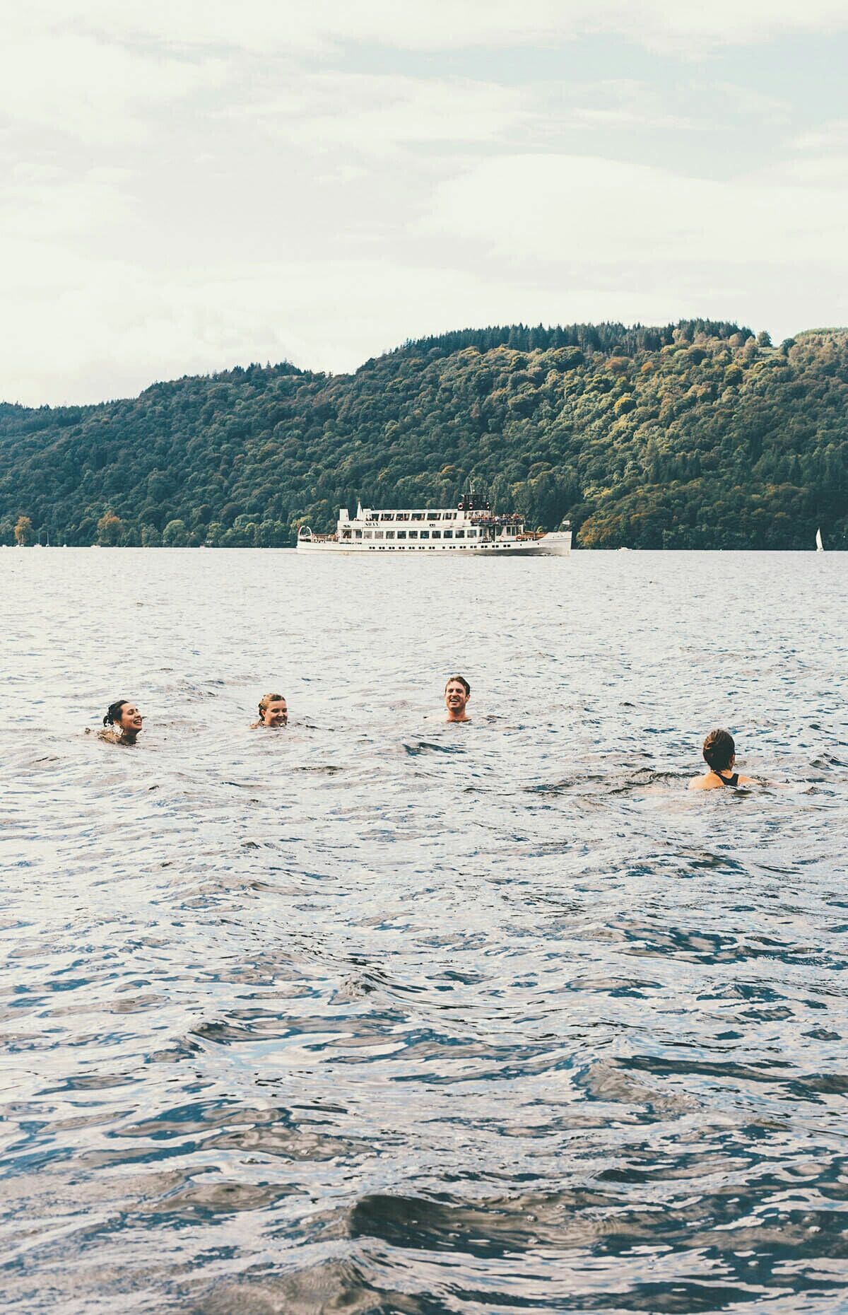 A group of people swimming in a lake with a boat and forested hill in the background.