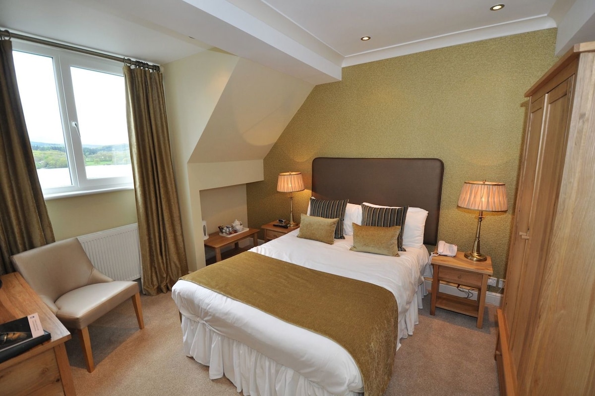 Habitación de hotel con una cama grande, dos mesitas de noche con lámparas, una silla, un escritorio y vistas al exterior.