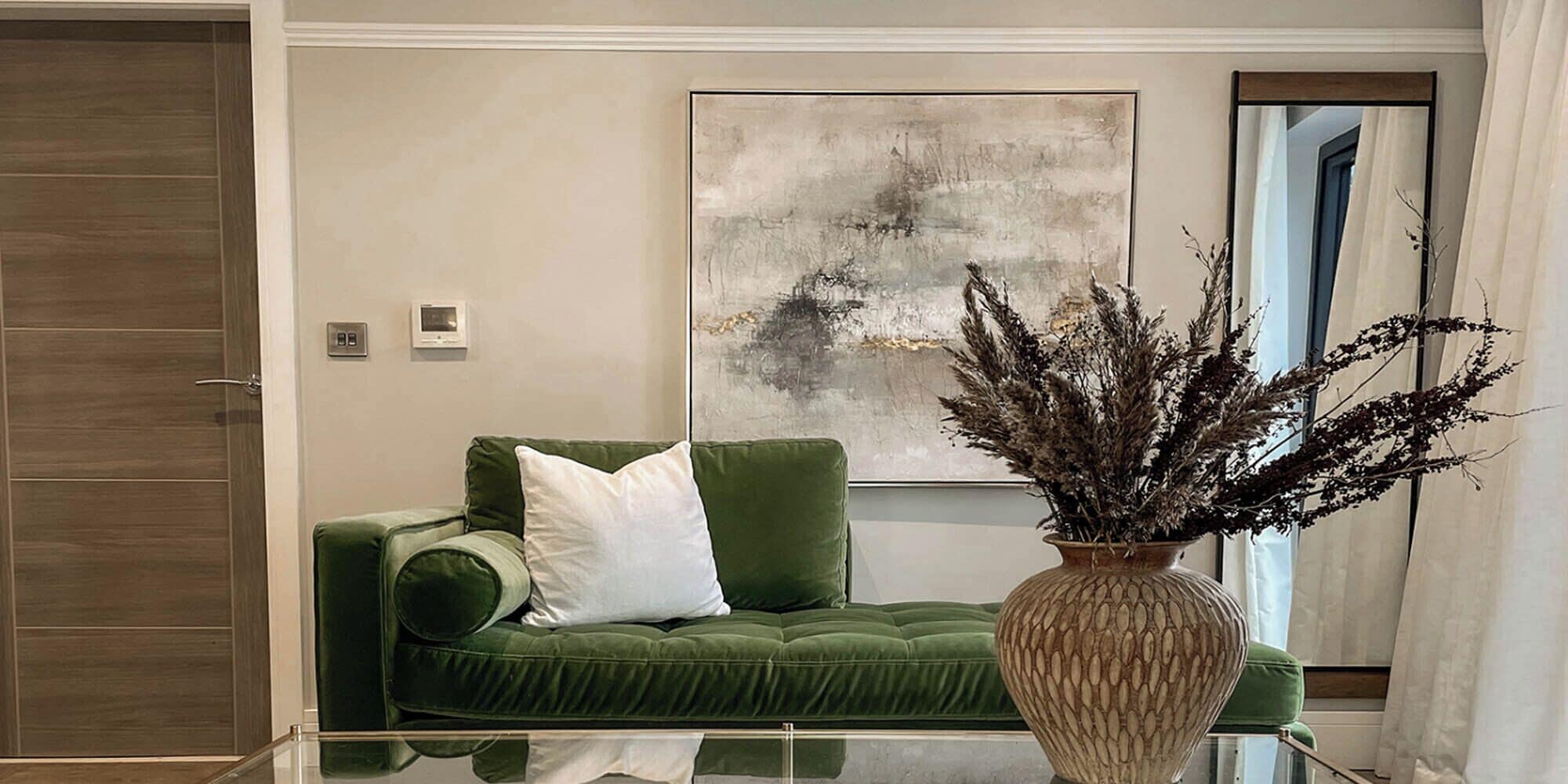 A green sofa with white cushions, a decorative vase with dried plants, and a large abstract painting on the wall.