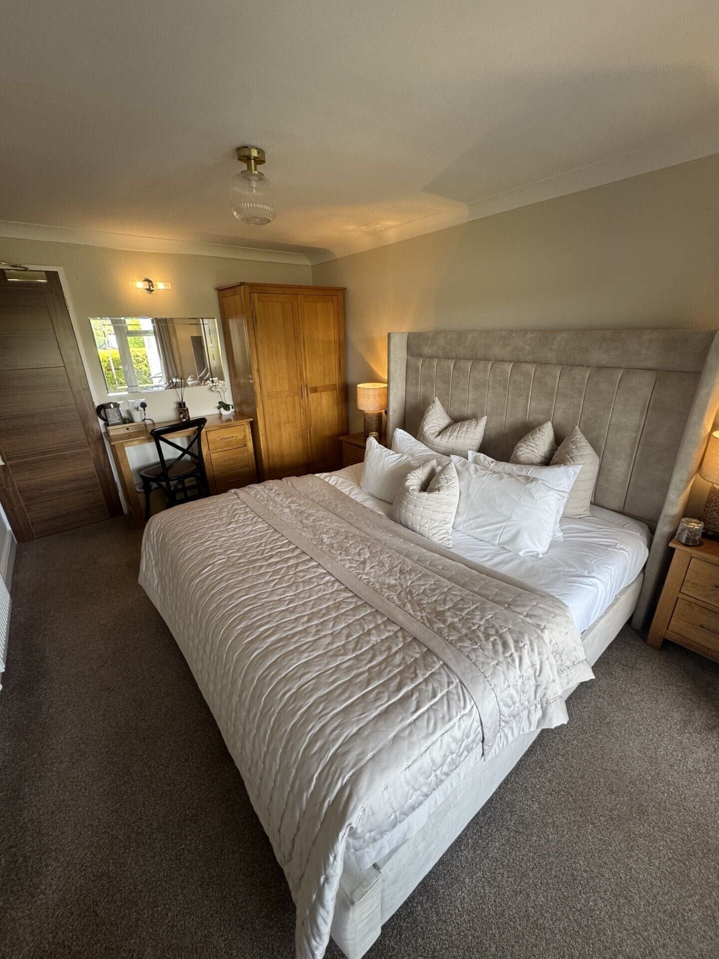 A bedroom with a large bed, a wooden wardrobe, a desk with a chair, and a mirror.