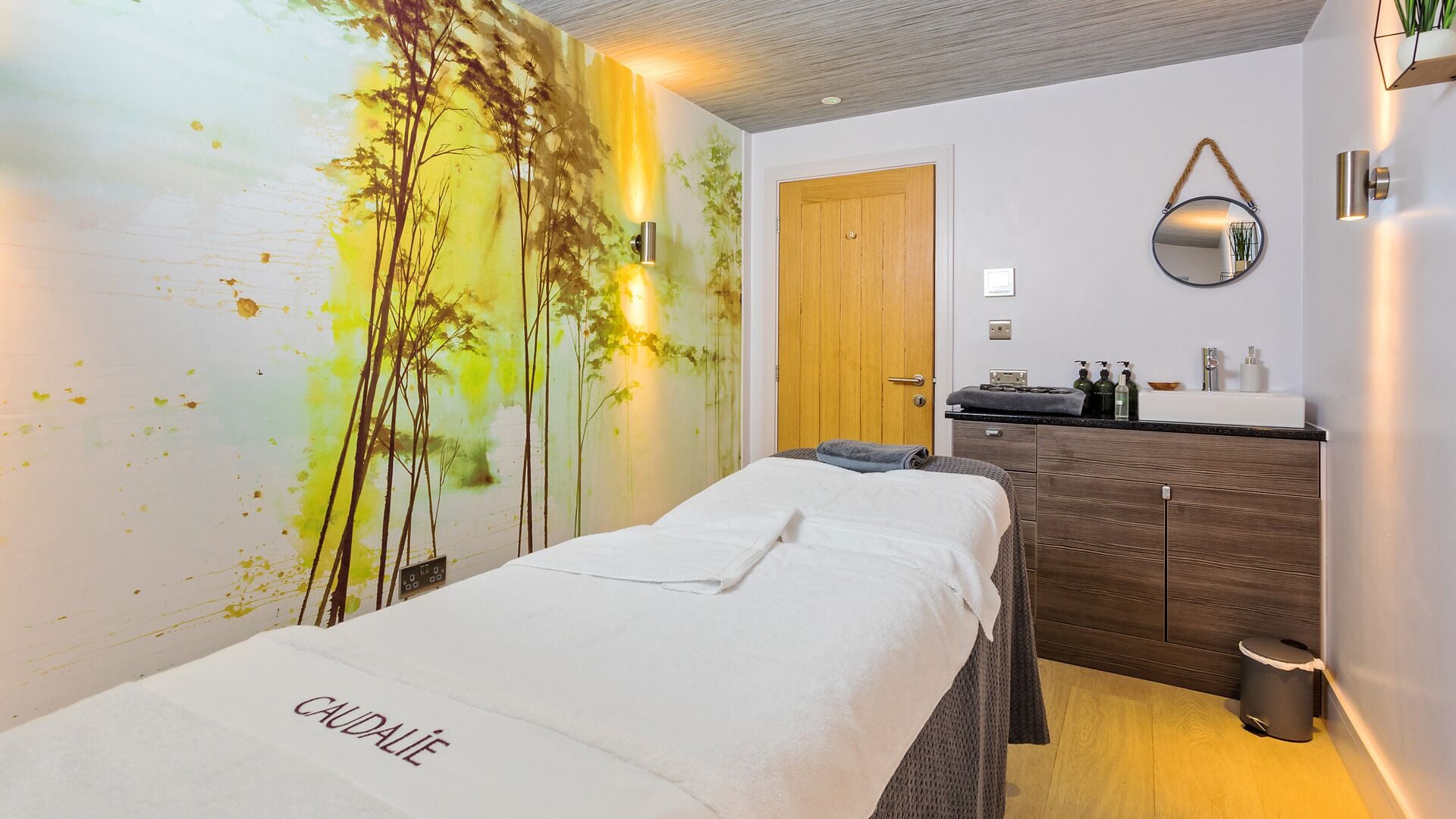 A spa room with a bed labeled 'CAUDALIE', a wooden door, a mirror, a sink with toiletries, and a wall with a nature-themed mural.