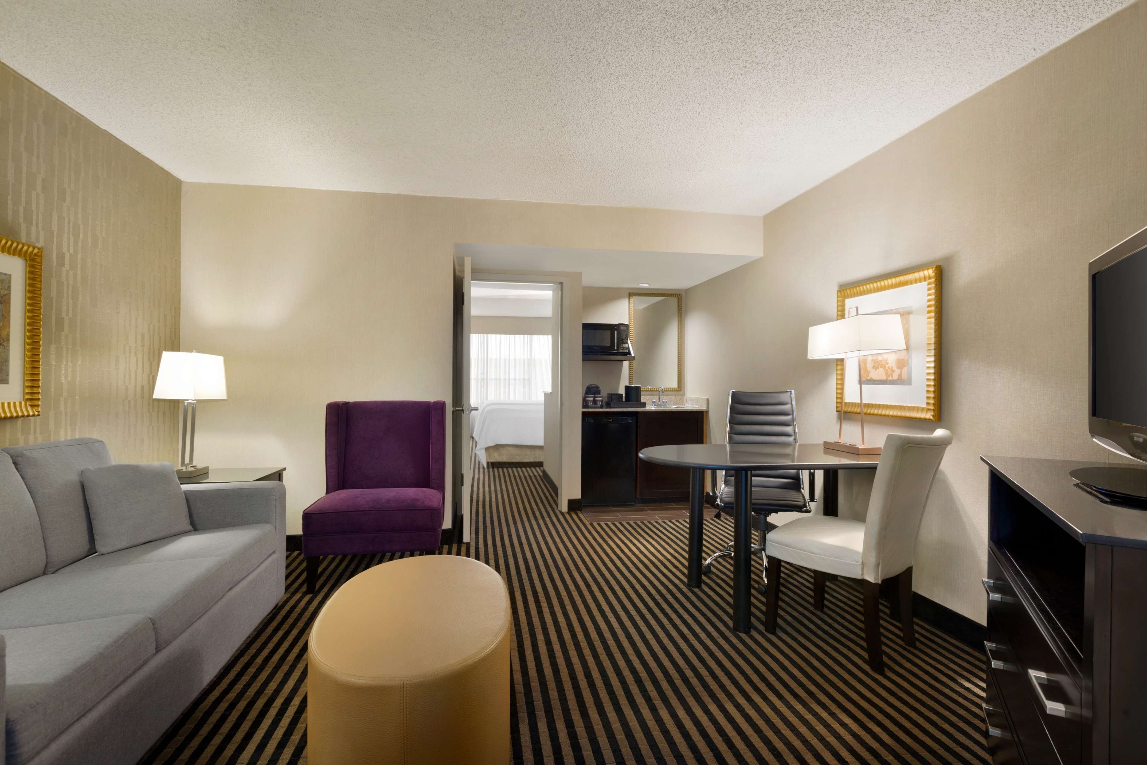 A hotel room with a sofa, a purple armchair, a small round table, a dining table with chairs, a TV, and a kitchenette.