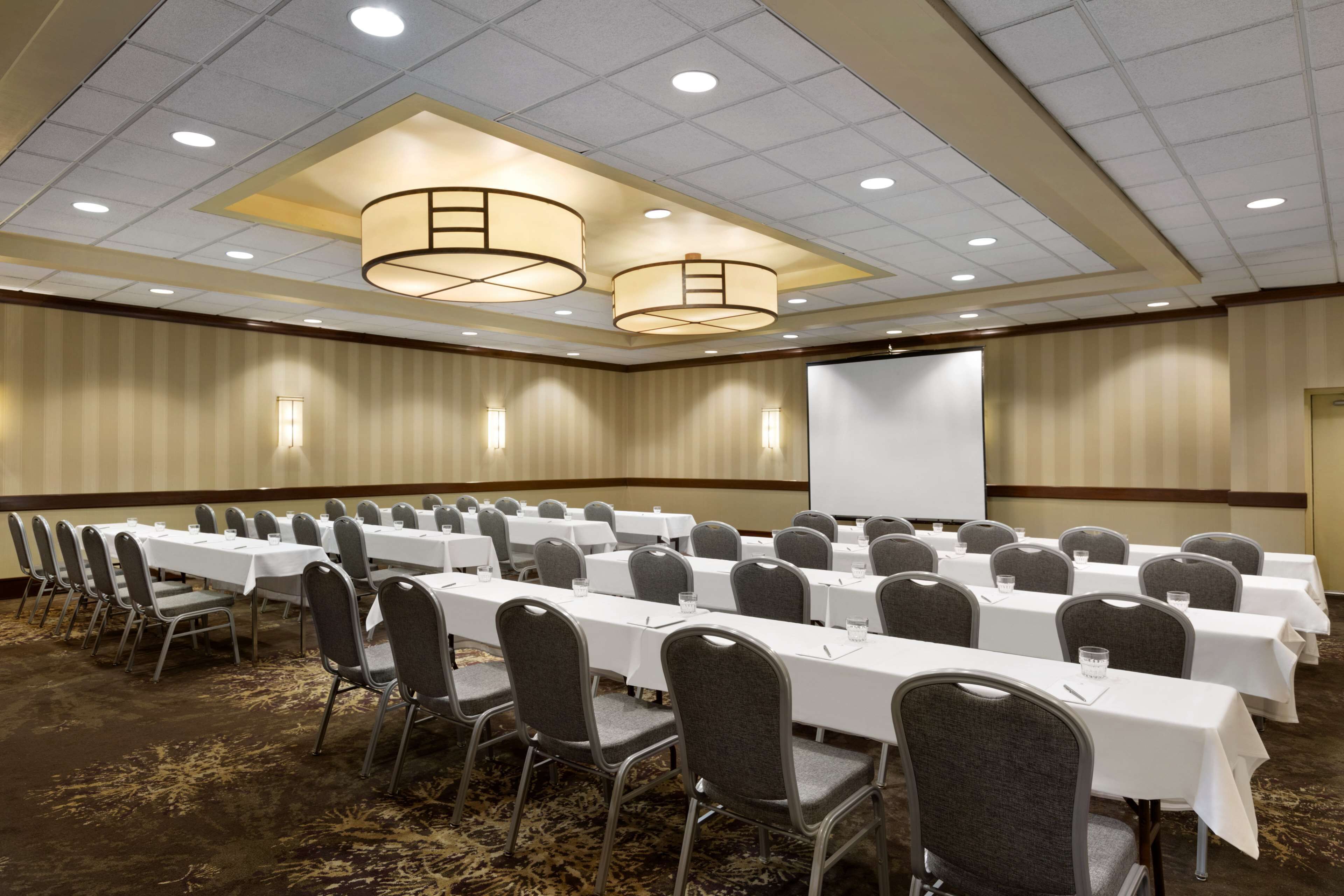 A conference room with rows of tables and chairs, a projector screen, and a carpeted floor.