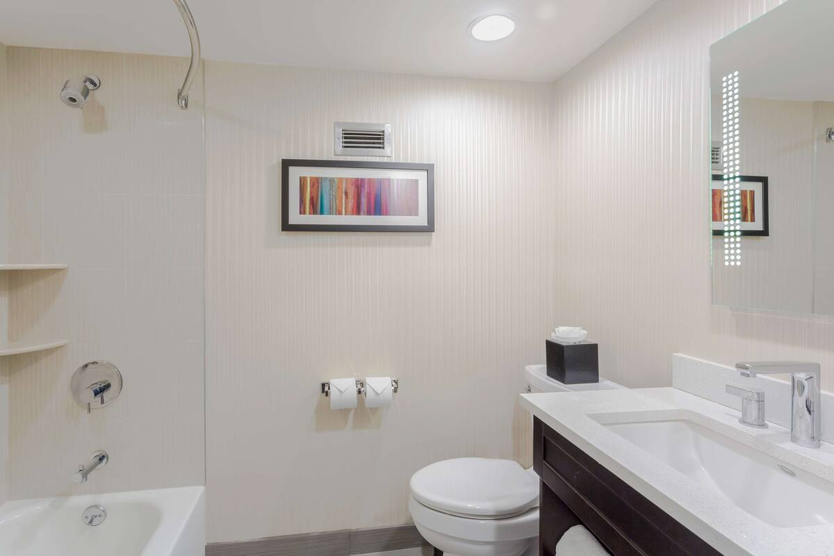 A modern bathroom with a bathtub, toilet, and sink. There is a framed artwork on the wall and a mirror with lights.