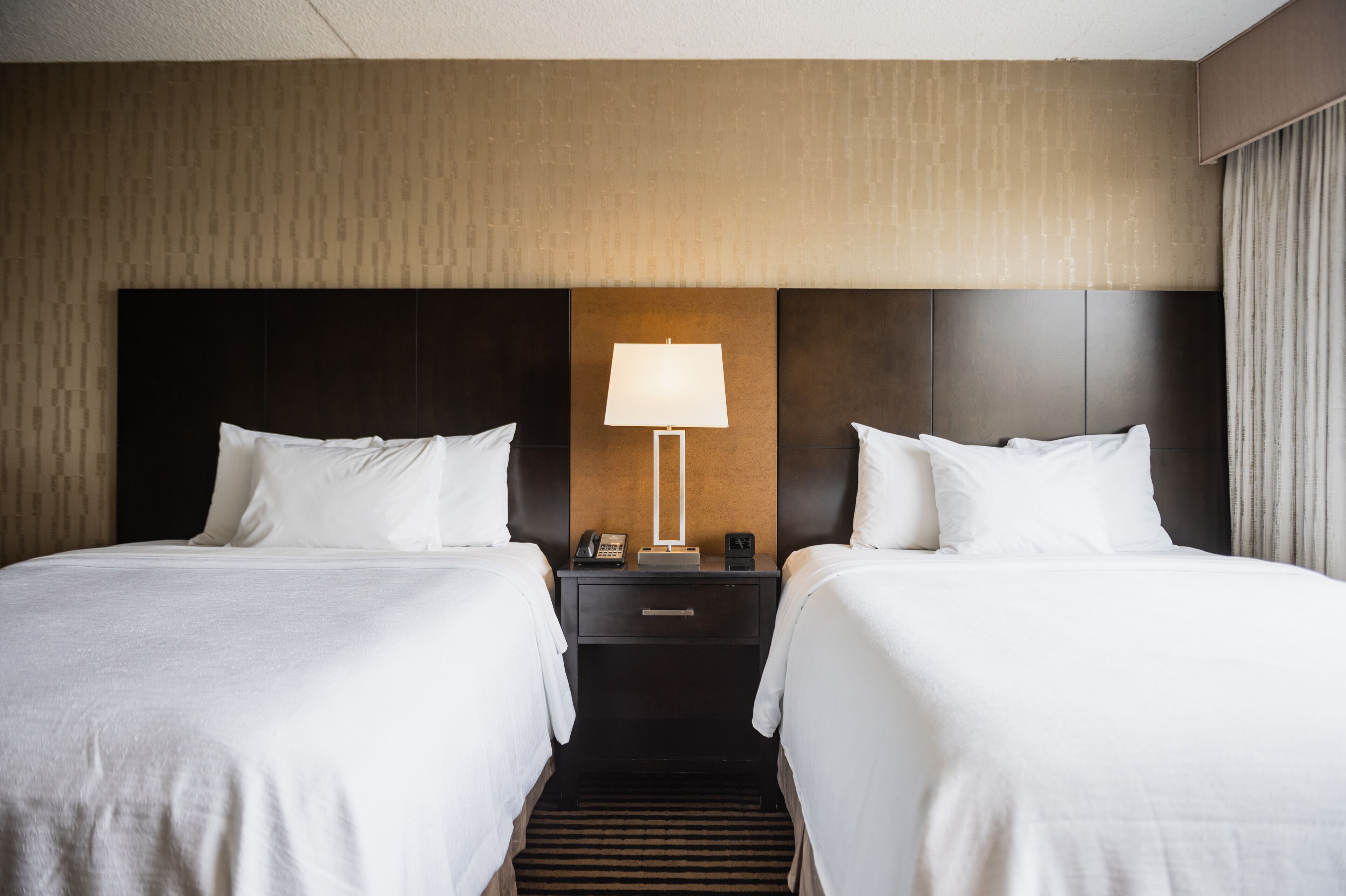A hotel room with two beds, a nightstand with a lamp, and a wall-mounted phone.